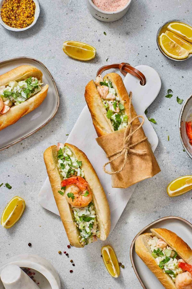 Overhead view of New England shrimp rolls with herbs in baguette rolls, served on plates and a cutting board with lemon wedges and condiments on the side.
