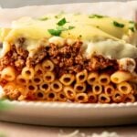 A close-up of a slice of pastitsio with ground lamb, showing layers of pasta, meat sauce, and creamy topping, garnished with herbs. Text overlay details recipe information.