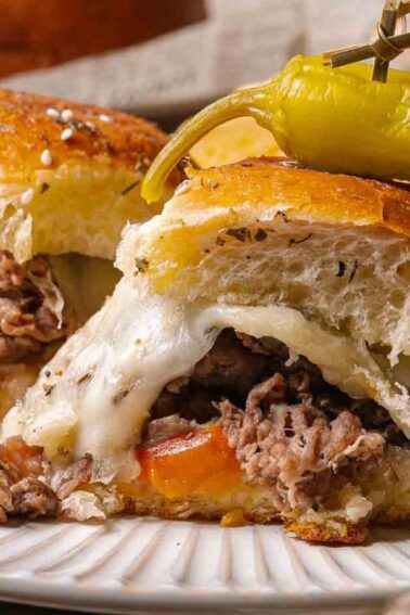 A plate with a cheesesteak sliders filled with melted cheese, sliced beef, peppers and onions, topped with a pepper, served alongside potato chips.