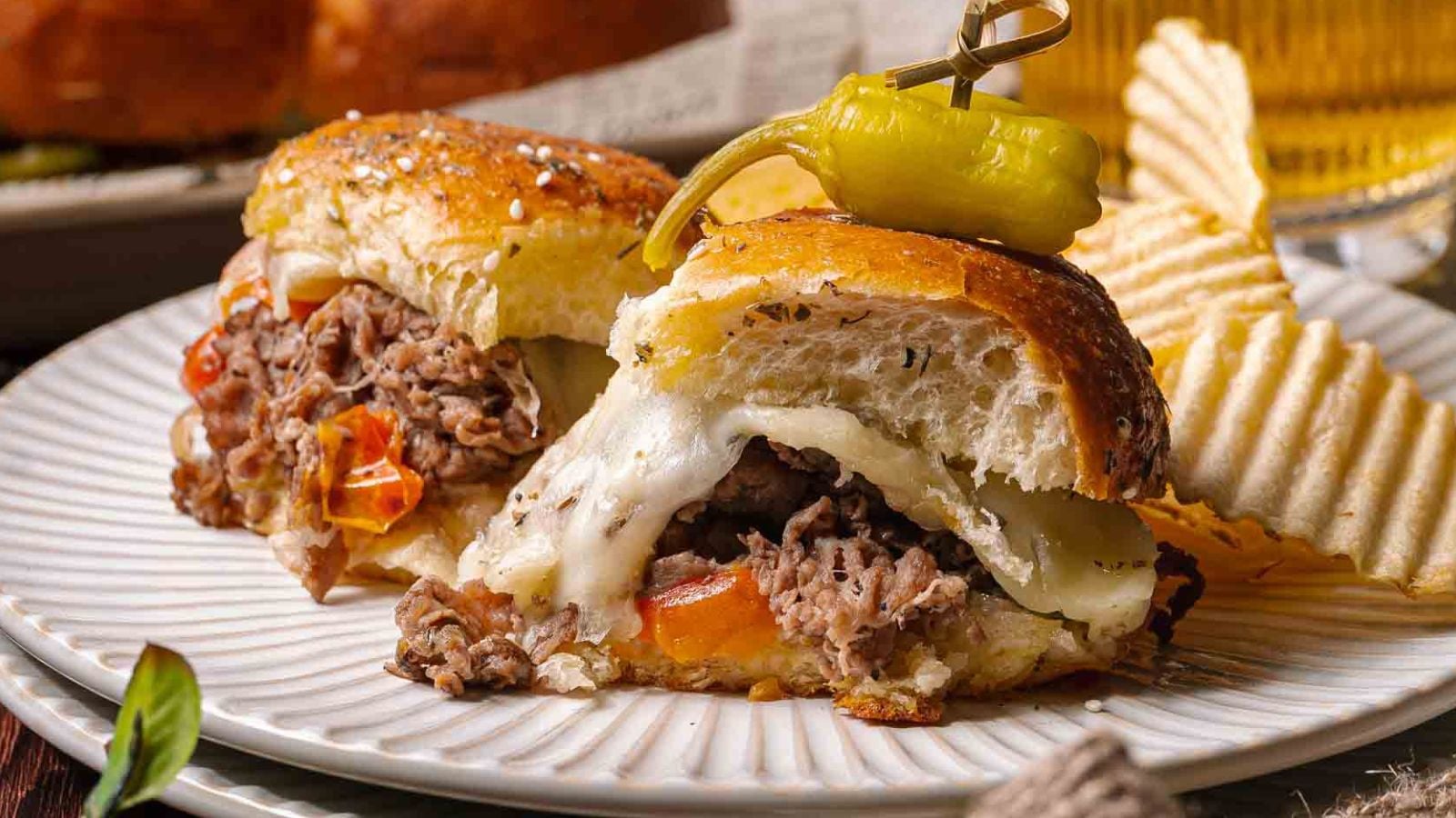 A plate with a cheesesteak sliders filled with melted cheese, sliced beef, peppers and onions, topped with a pepper, served alongside potato chips.