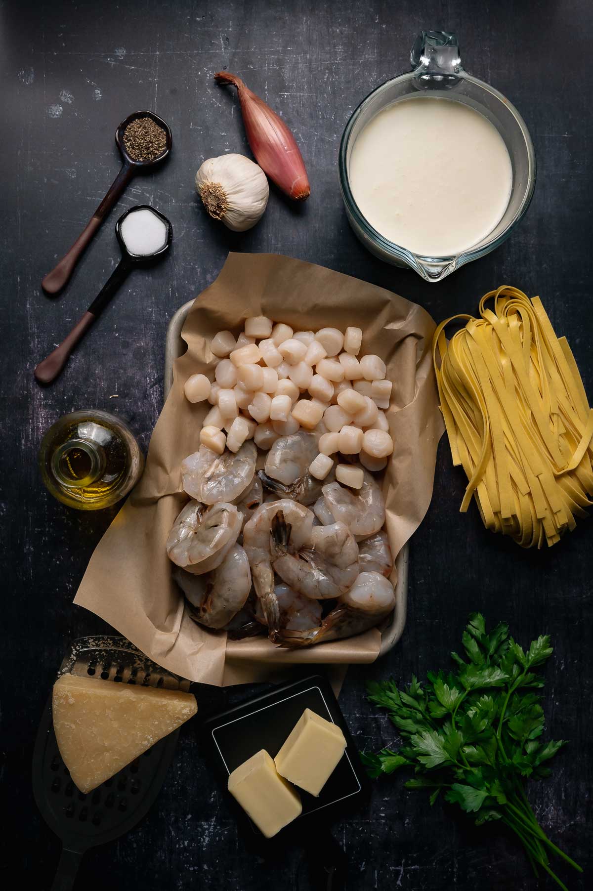 Ingredients for a seafood alfredo dish are arranged on a dark surface, including shrimp, scallops, fettuccine, cream, parmesan, butter, parsley, shallot, garlic, olive oil, salt, and pepper.