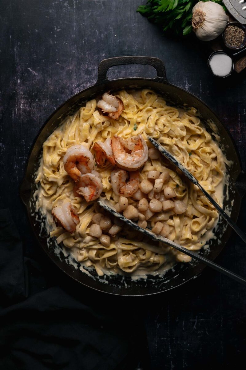 A skillet filled with fettuccine Alfredo topped with shrimp and scallops, with metal tongs resting on top; garlic, parsley, and seasonings are nearby.