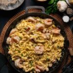 A skillet filled with fettuccine pasta and shrimp in a creamy alfredo sauce, garnished with herbs, with bread, garlic, parmesan, salt, and pepper on the side.