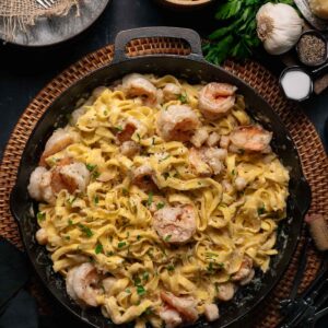 A skillet filled with fettuccine pasta and shrimp in a creamy alfredo sauce, garnished with herbs, with bread, garlic, parmesan, salt, and pepper on the side.