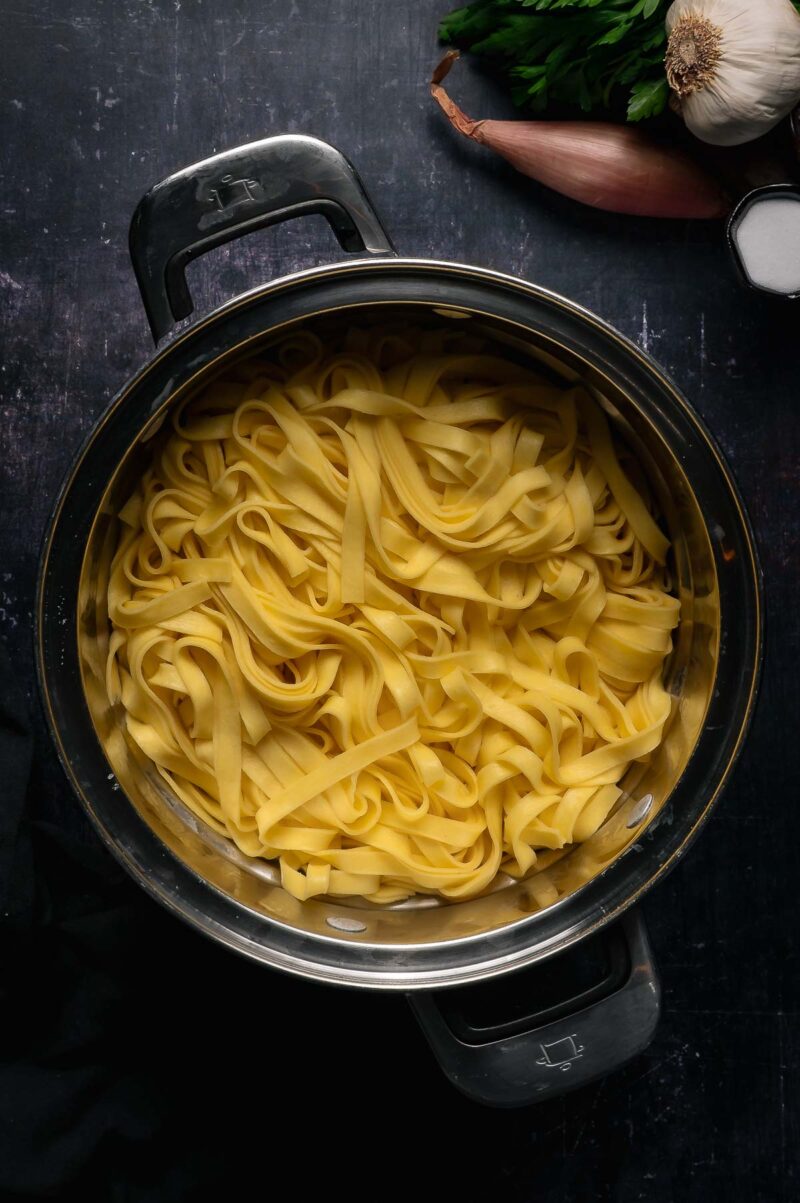 A pot of cooked fettuccine pasta sits on a dark surface, with garlic, parsley, a shallot, and a bowl of salt nearby.