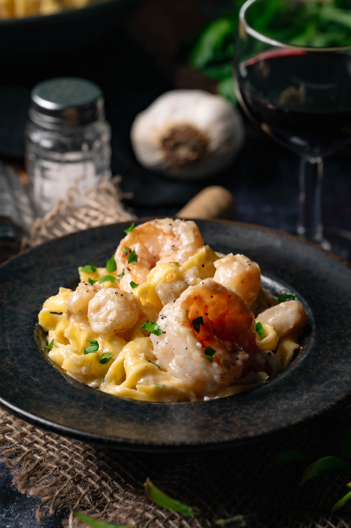 A plate of seafood fettuccine alfredo pasta in a creamy sauce, garnished with herbs, is served next to a glass of red wine and a head of garlic.