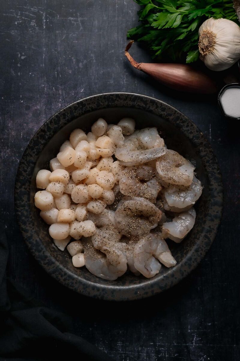 A bowl containing raw scallops and shrimp seasoned with pepper, next to fresh parsley, a shallot, a garlic bulb, and a small bowl of salt.