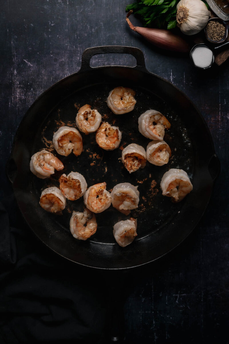 A black skillet with shrimp arranged in a circle, with garlic, shallot, parsley, salt, and pepper visible in the top right corner of the image.