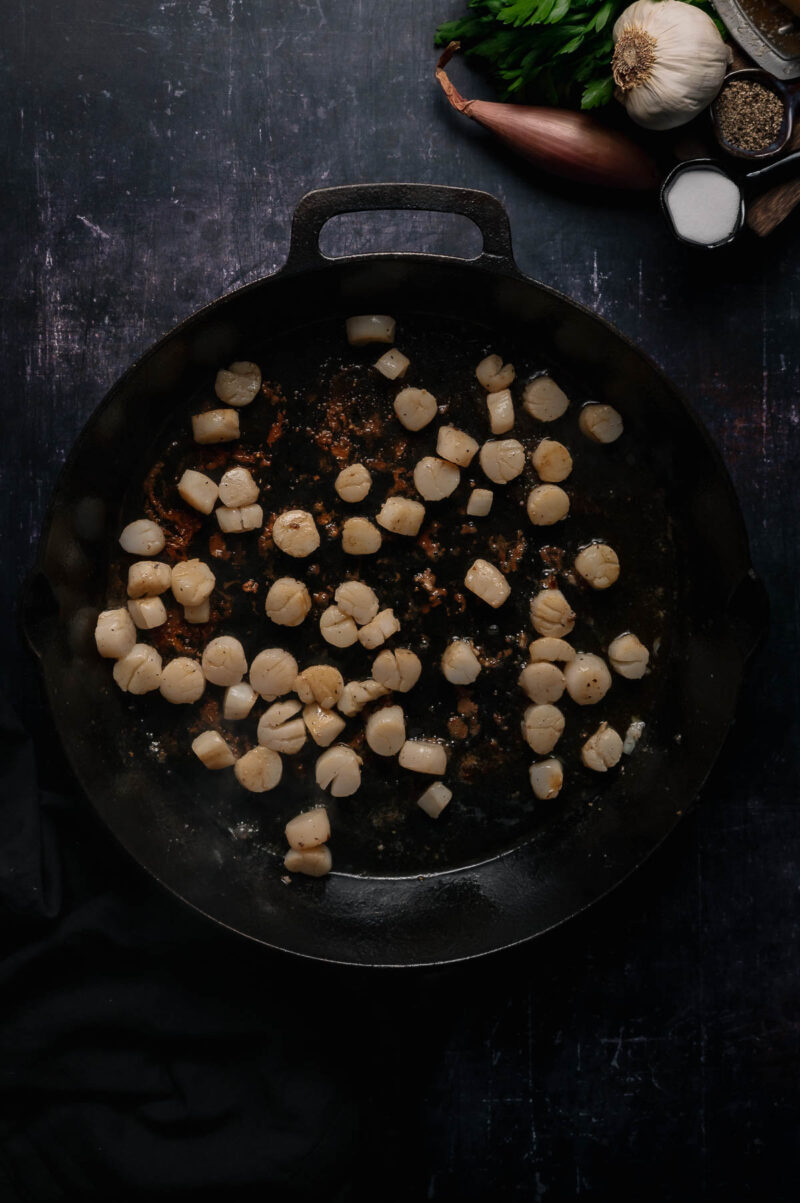 Scallops cooking in a black cast iron pan, surrounded by garlic, parsley, shallot, salt, pepper, and a cloth on a dark surface.