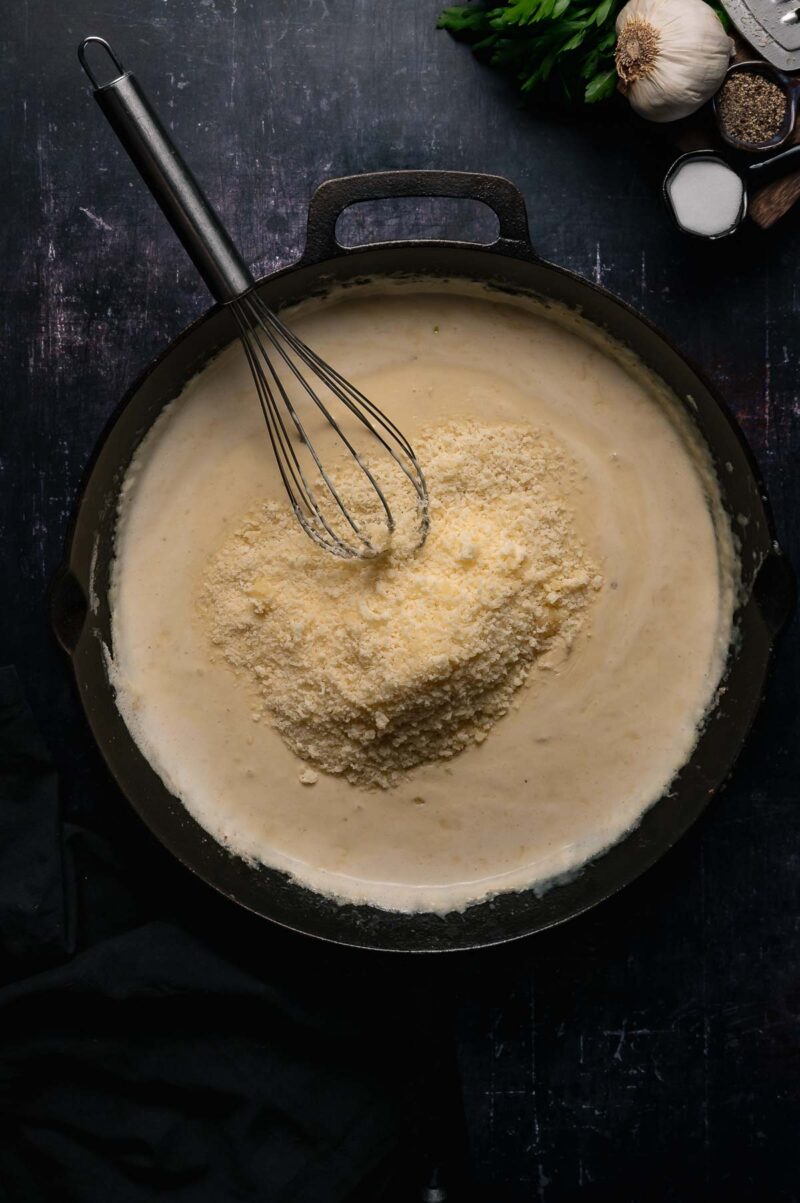A whisk rests in a pan of creamy alfredo sauce with a mound of grated cheese on top; garlic, salt, and parsley are nearby.