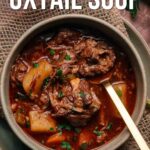 A bowl of slow cooker oxtail soup with chunks of meat and vegetables, garnished with chopped herbs, sits on a burlap mat with a spoon inside.