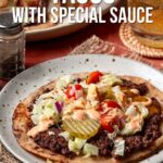 A plate with a smash burger taco topped with lettuce, pickles, tomatoes, cheese, and special sauce. A glass of drink and condiments are in the background.