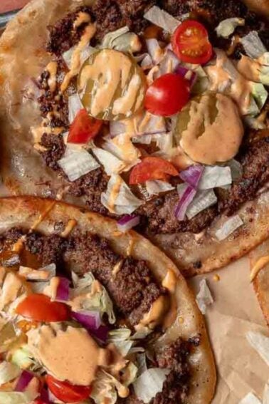 Three flatbread pizzas topped with ground meat, lettuce, tomato, pickles, onions, and a drizzle of sauce are arranged on a baking tray next to a small bowl of pickle slices.