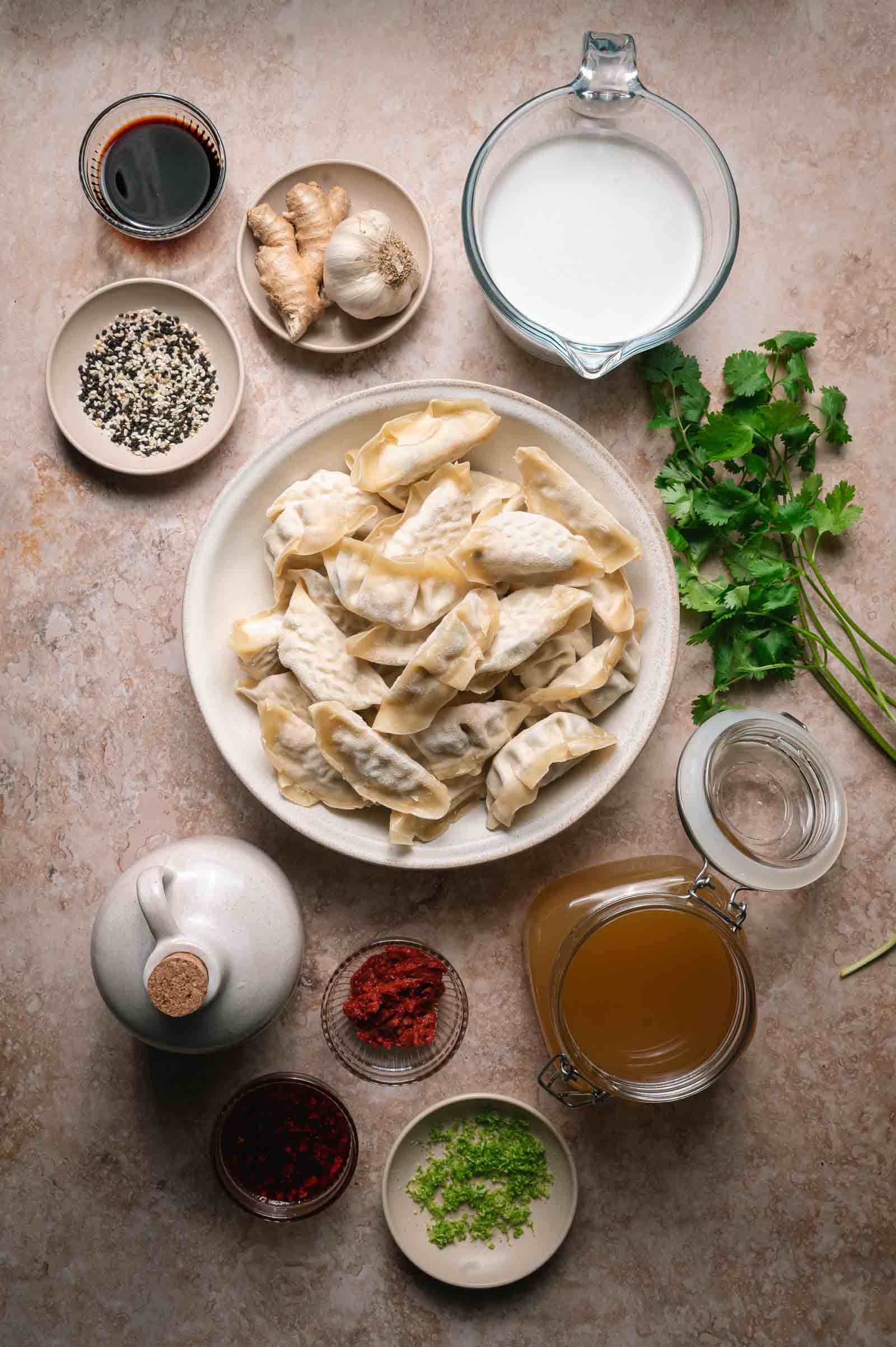 A bowl of uncooked dumplings surrounded by small dishes of sauces, spices, fresh herbs, ginger, garlic, and a glass pitcher of broth on a countertop.