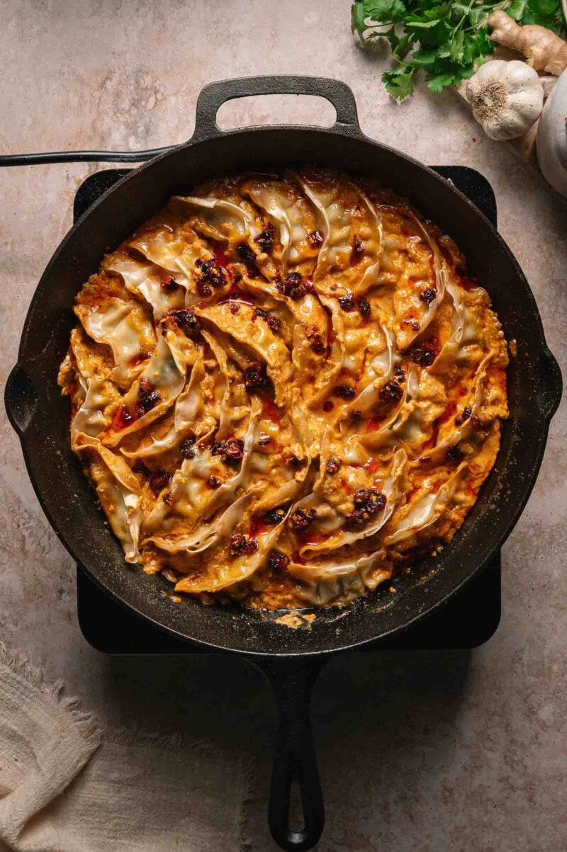 Cast iron skillet filled with cooked dumplings in a creamy coconut sauce, garnished with chili oil, placed on a light stone surface.