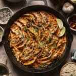 A cast iron skillet filled with arranged pan-fried dumplings in creamy coconut sauce, garnished with herbs, sesame seeds, and lime wedges, surrounded by rice, sauces, and chopsticks.