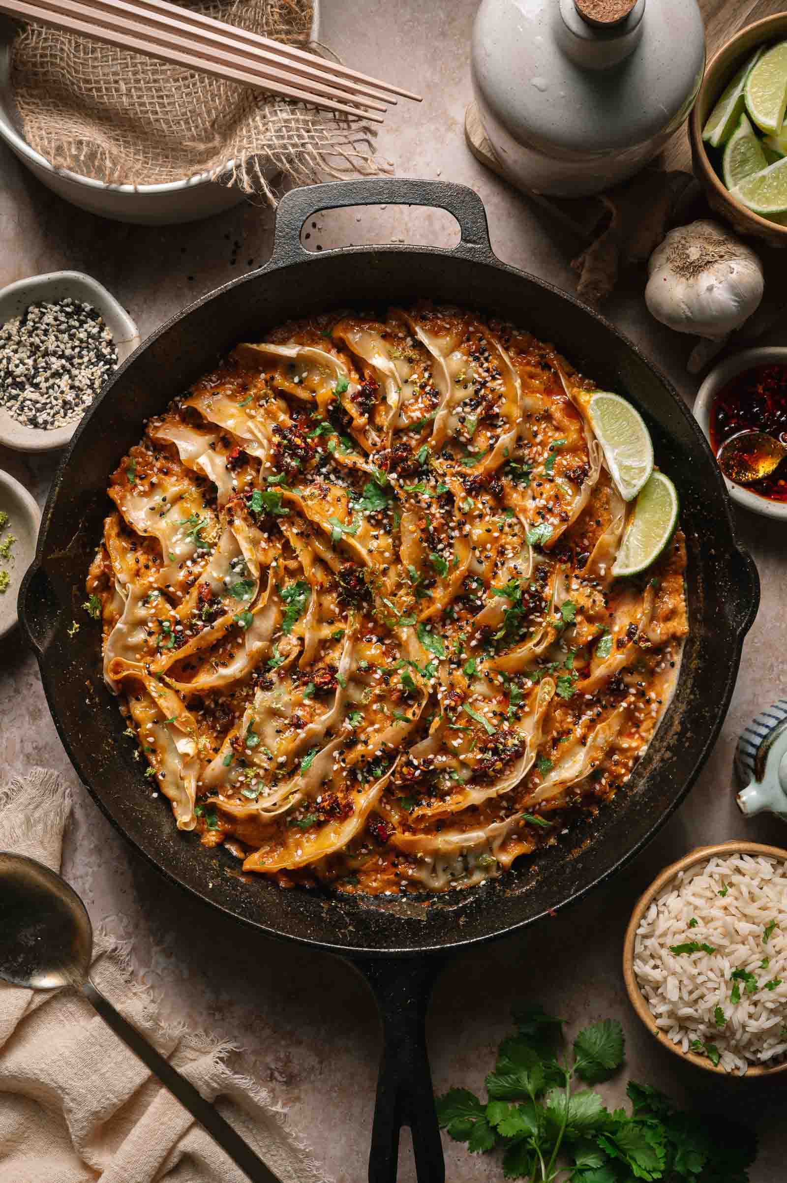 A cast iron skillet filled with arranged pan-fried dumplings in creamy coconut sauce, garnished with herbs, sesame seeds, and lime wedges, surrounded by rice, sauces, and chopsticks.
