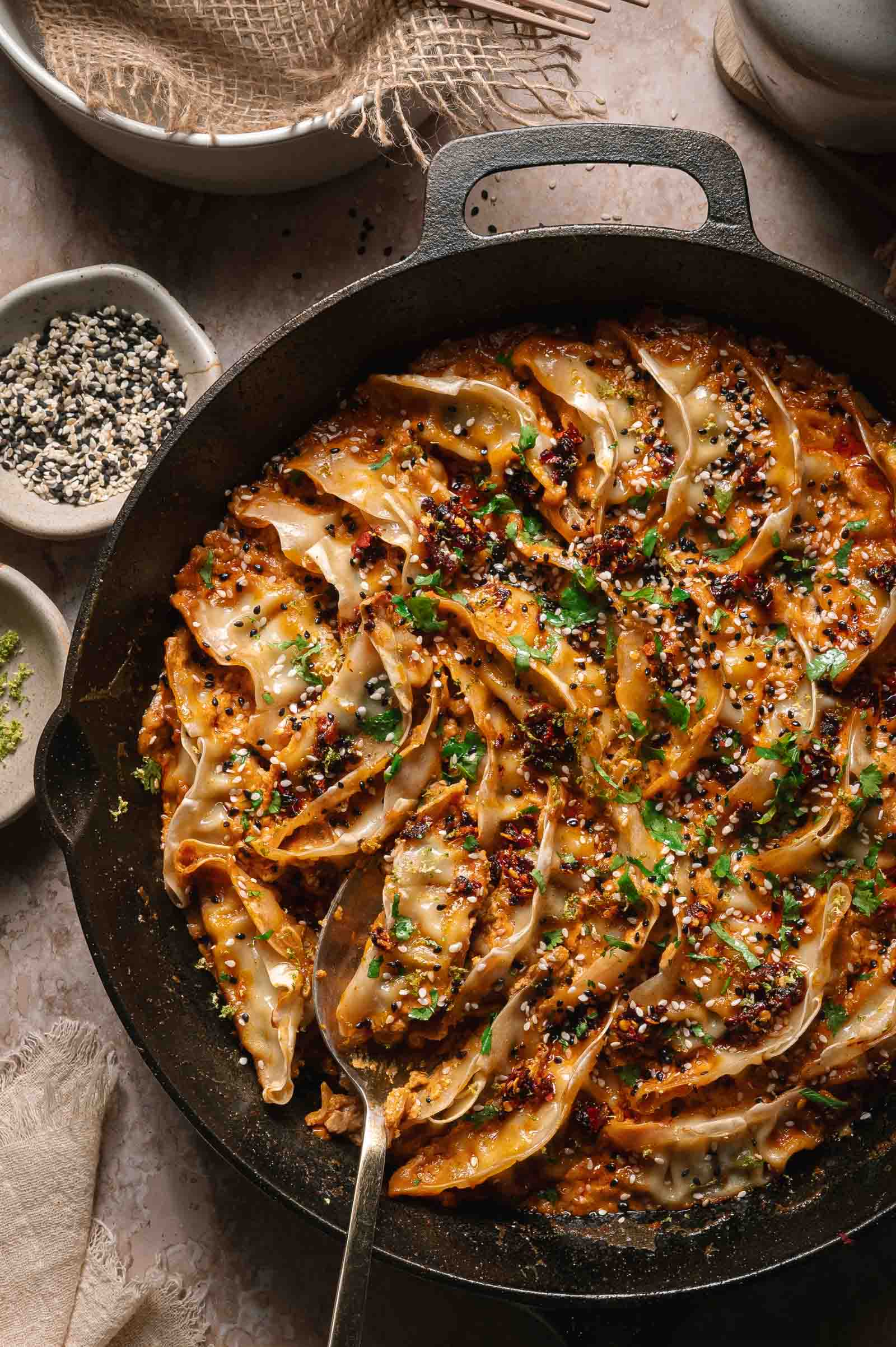 A cast iron skillet filled with arranged dumplings topped with sauce, lime zest, and sesame seeds, with a spoon and small bowls of seasonings on the side.