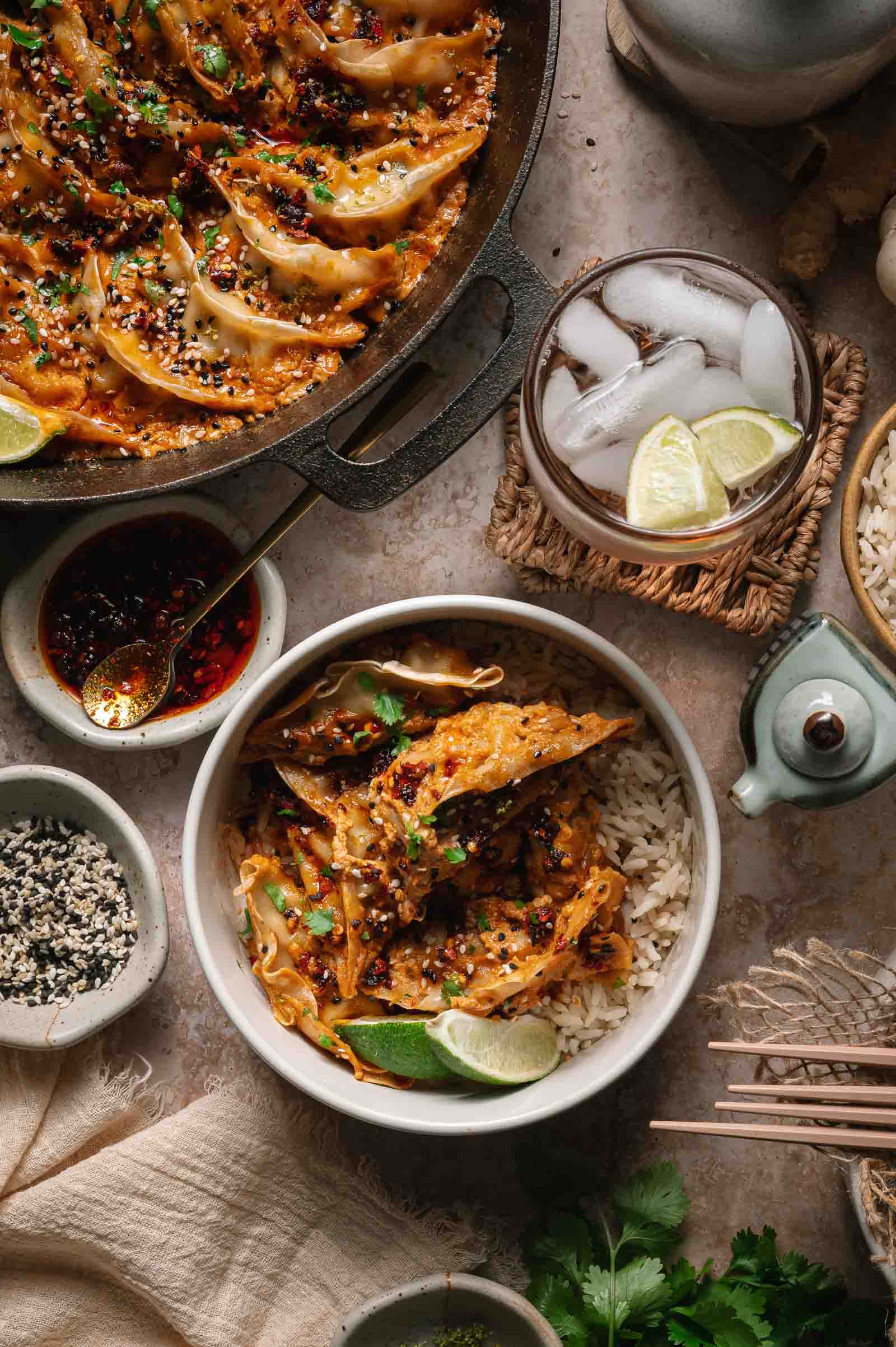 A bowl of rice topped with saucy baked dumplings, garnished with sesame seeds, cilantro, lime wedges, and chili oil, surrounded by a pan of dumplings, rice, and a glass of iced water.