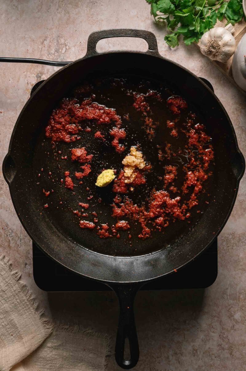 A cast iron skillet on a stovetop with red curry paste, minced ginger, and garlic; fresh cilantro and garlic are beside the skillet.