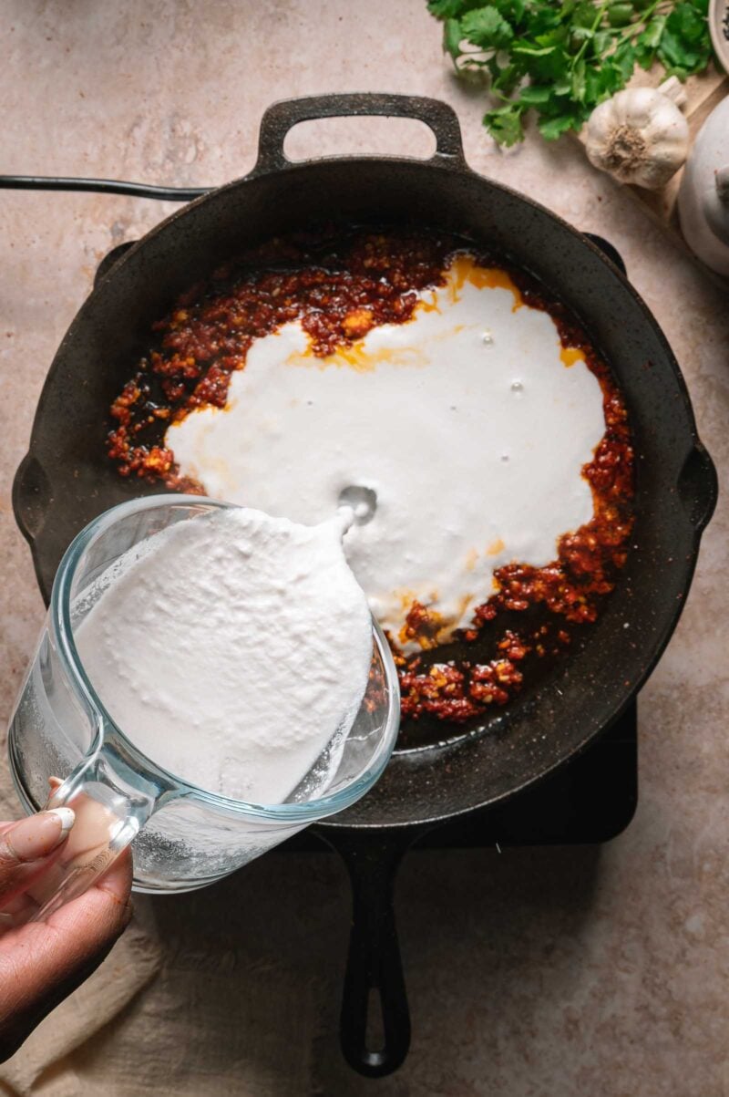 A hand pours coconut milk from a glass measuring cup into a cast iron skillet with a red curry paste mixture, with herbs and garlic nearby.