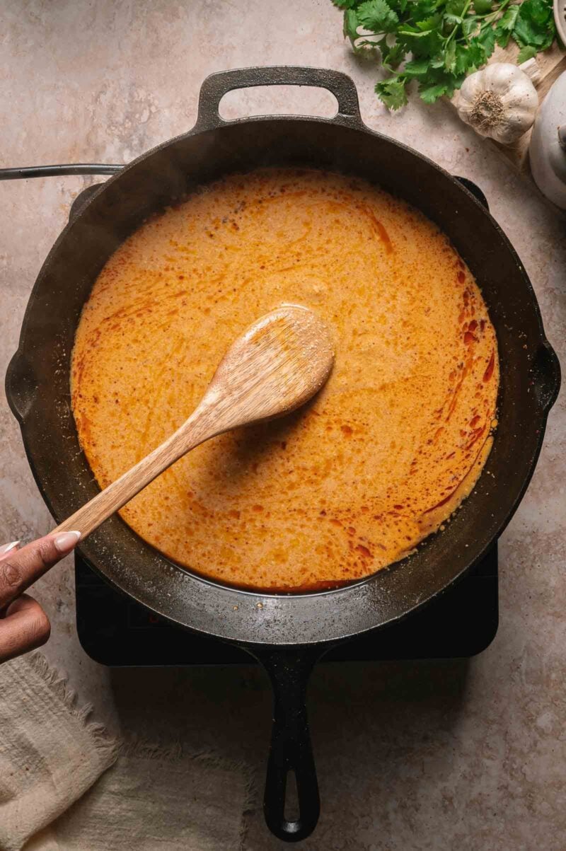 A person stirs a creamy, coconut curry sauce in a black cast iron skillet with a wooden spoon; herbs and garlic are nearby.
