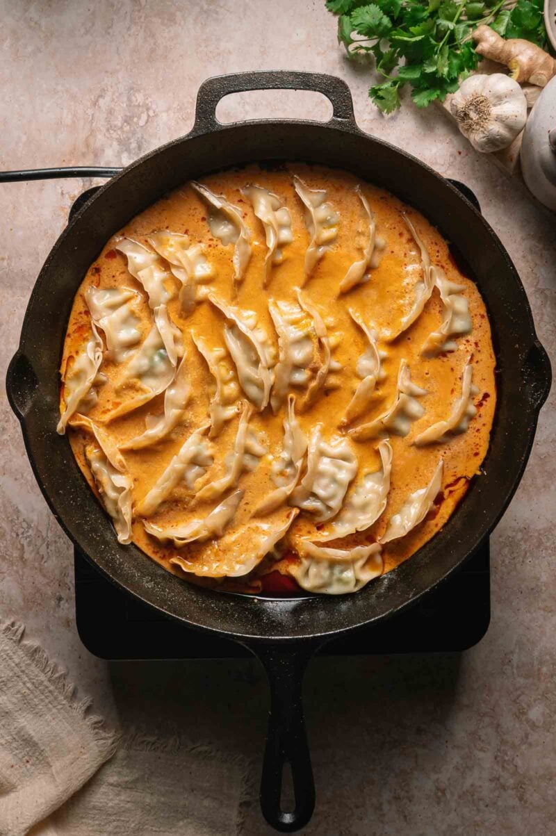 A cast iron skillet filled with dumplings arranged in a creamy coconut curry sauce, with fresh herbs and garlic visible nearby on a light kitchen surface.