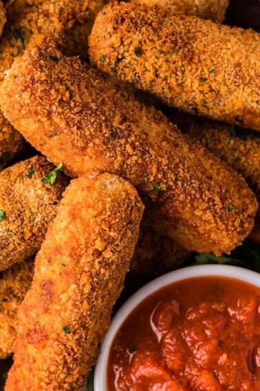 A pile of breaded mozzarella sticks arranged on a platter with a bowl of marinara sauce for dipping, garnished with parsley.