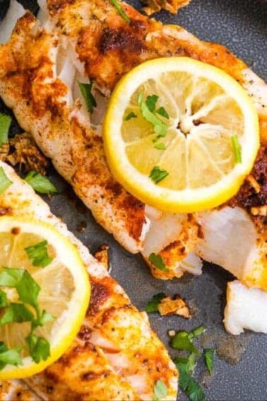 Two pieces of seasoned, cooked white fish fillets topped with lemon slices and chopped herbs on a dark plate.