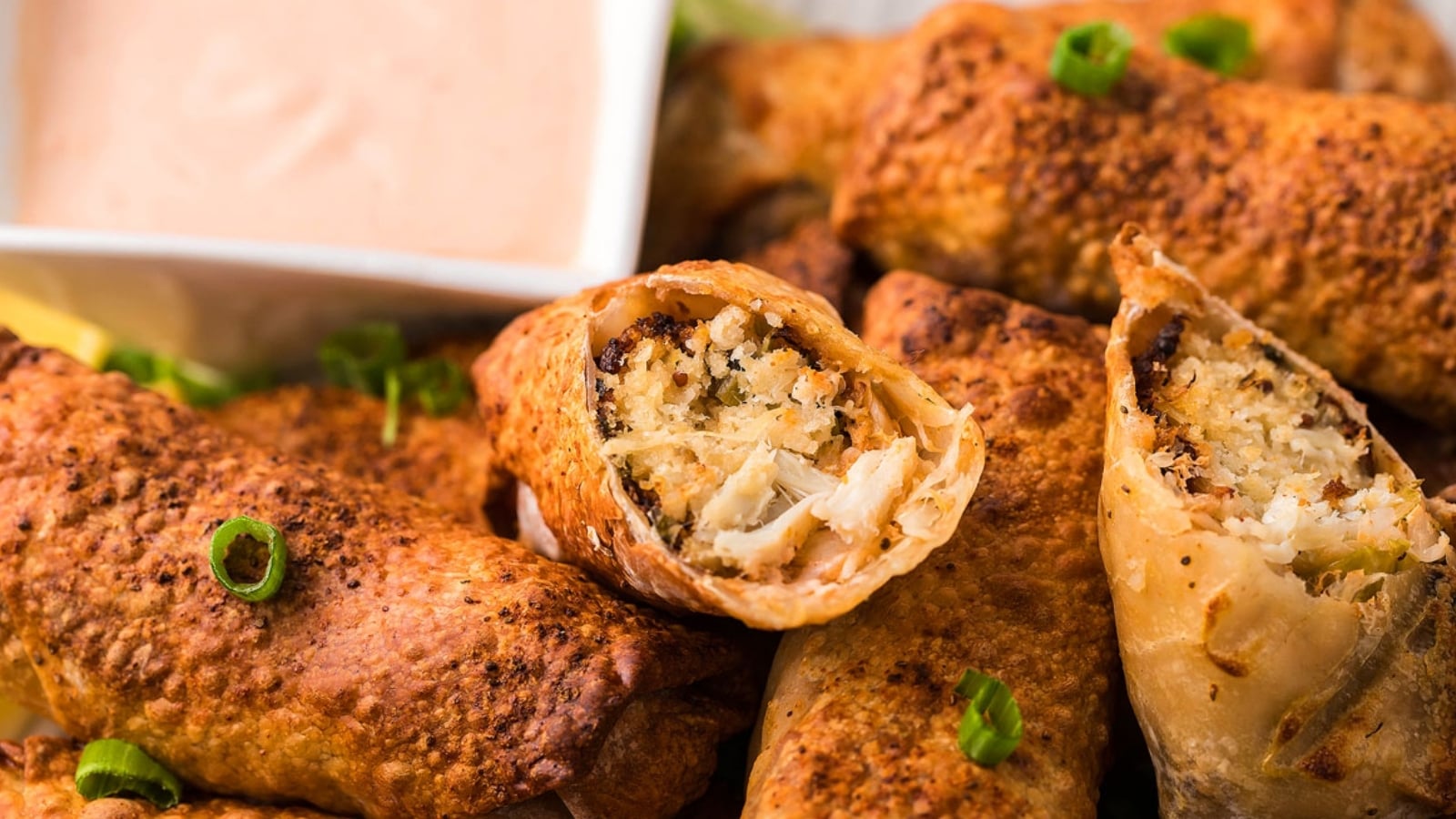 Close-up of crispy egg rolls filled with crab meat and rice, garnished with sliced green onions, with a small bowl of dipping sauce in the background.