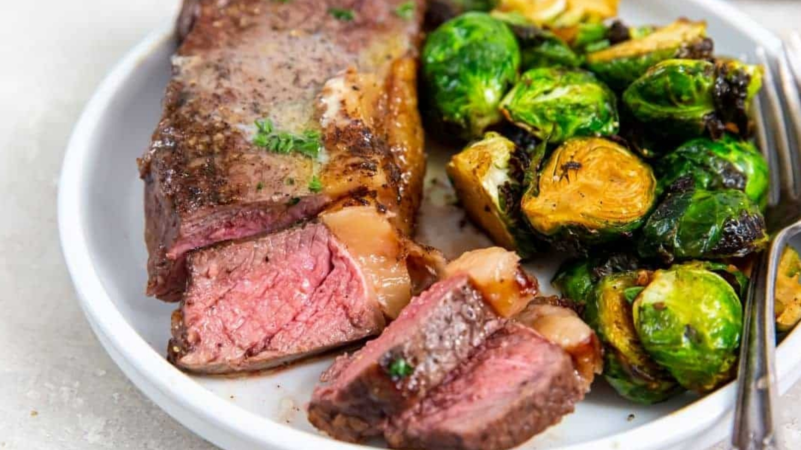 Air Fryer New York Strip Steaks on a white plate with butter, thyme, pepper, salt, brussels sprouts, and a fork.