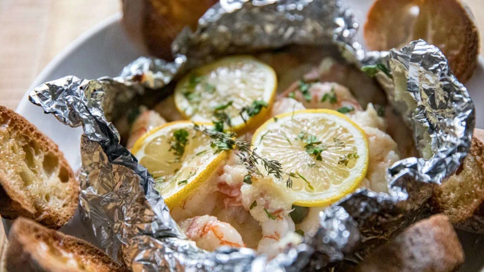 An opened foil packet filled with shrimp topped with lemon slices and chopped herbs sits in a bowl surrounded by toasted bread slices.