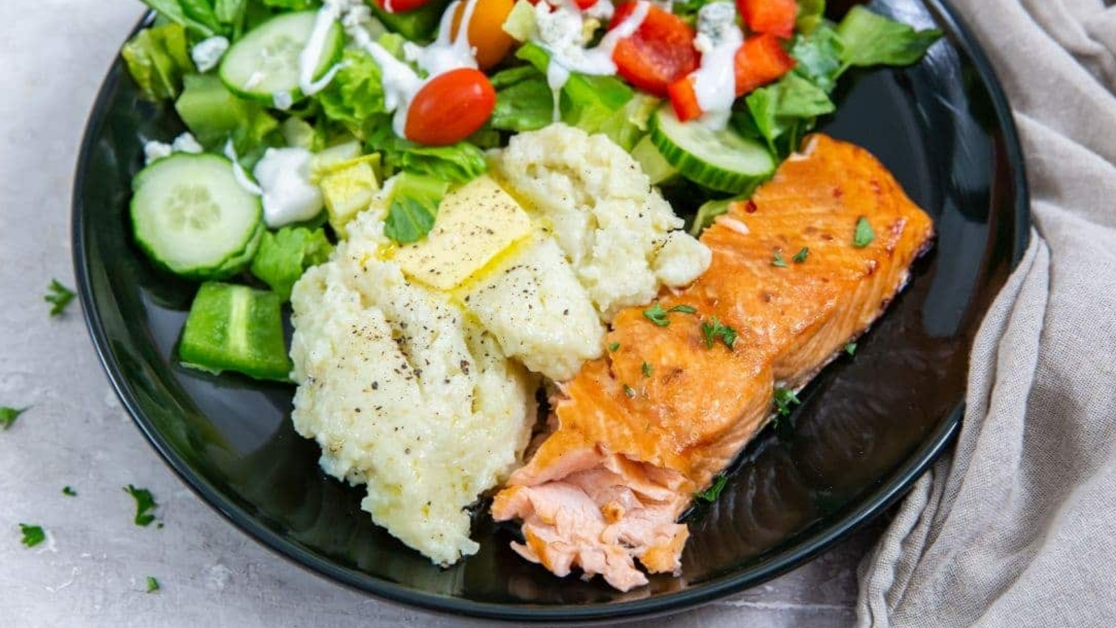 Air Fryer Teriyaki Salmon with rice, butter, parsley, black pepper, and a salad on a black plate.