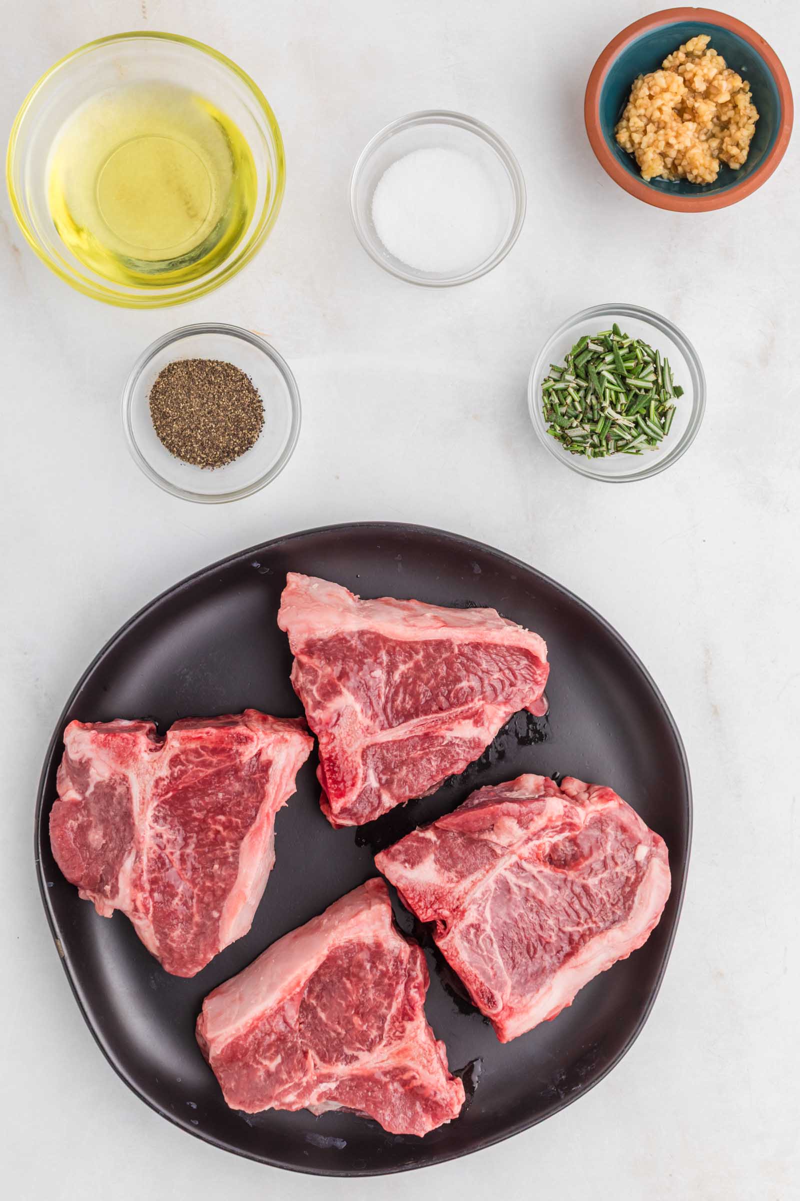 Four raw lamb loin chops on a black plate, surrounded by small bowls containing olive oil, salt, pepper, minced garlic, and chopped herbs on a white surface.