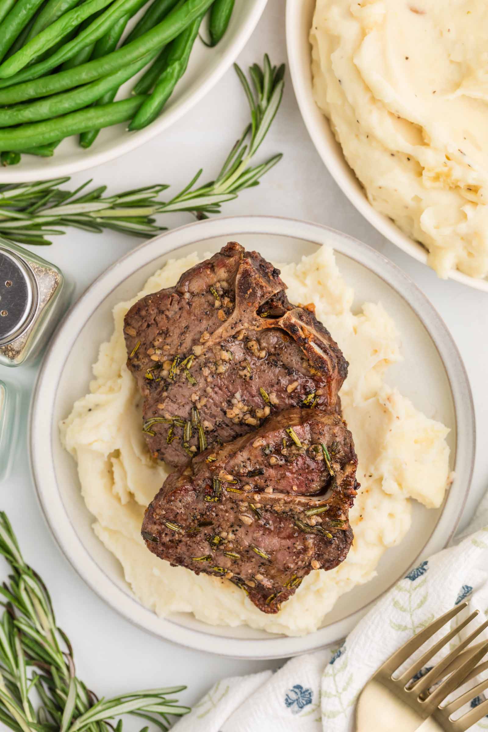Two cooked lamb chops with herbs are served on a bed of mashed potatoes. Green beans and extra mashed potatoes are in bowls nearby. A fork and sprigs of rosemary are also visible.