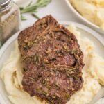 Grilled lamb chops with herbs served on a bed of mashed potatoes, with a bowl of green beans and a pepper shaker in the background.