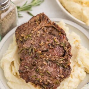 Grilled lamb chops with herbs served on a bed of mashed potatoes, with a bowl of green beans and a pepper shaker in the background.