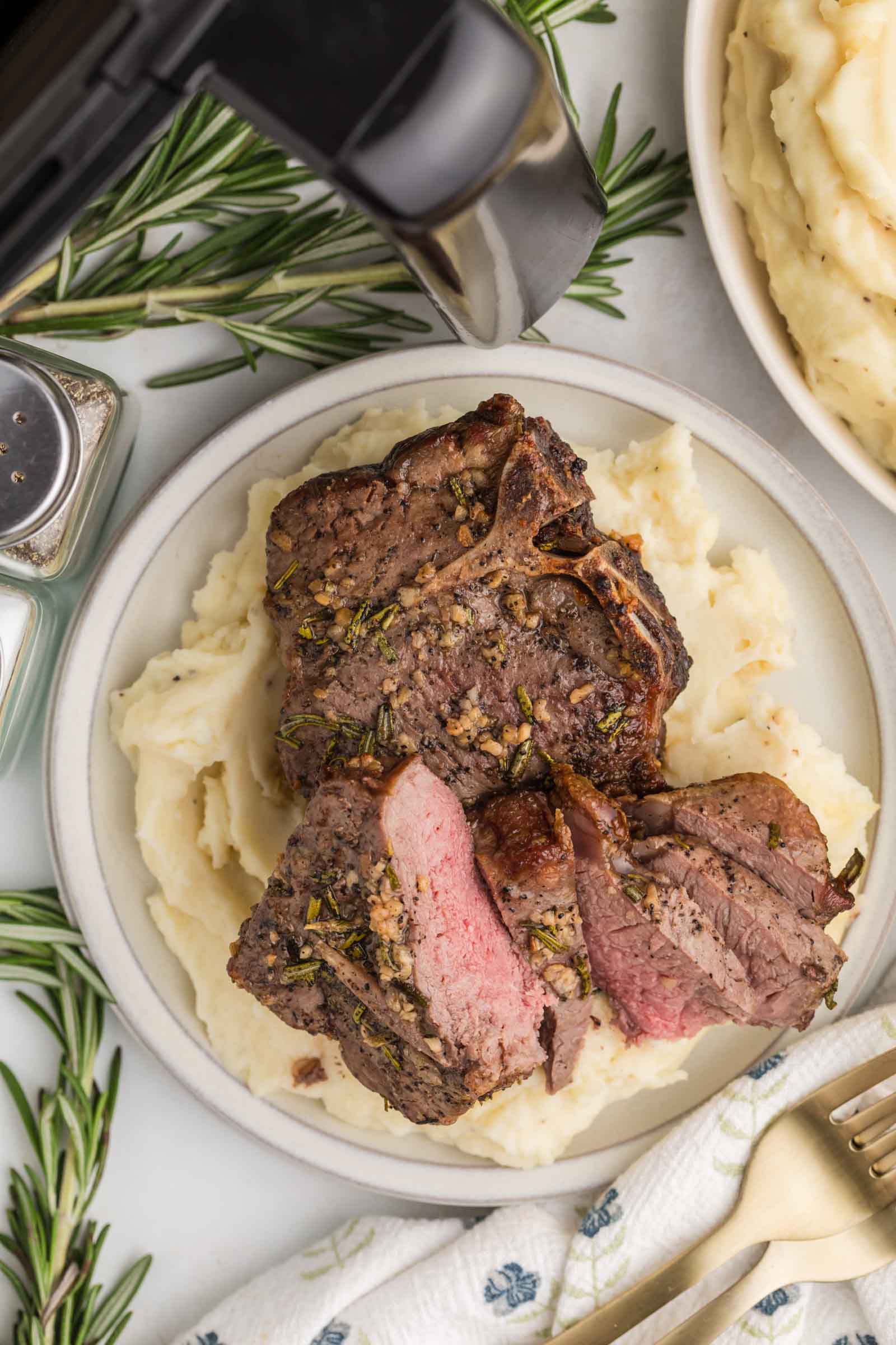 A plate of sliced lamb loin chops served on mashed potatoes, garnished with herbs, next to an air fryer, rosemary sprigs, and a bowl of mashed potatoes.