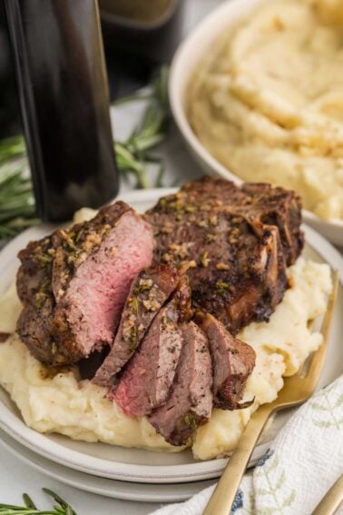 Sliced medium-rare lamb loin chops served on a bed of mashed potatoes, garnished with herbs, with a fork on the plate and a bowl of mashed potatoes in the background.