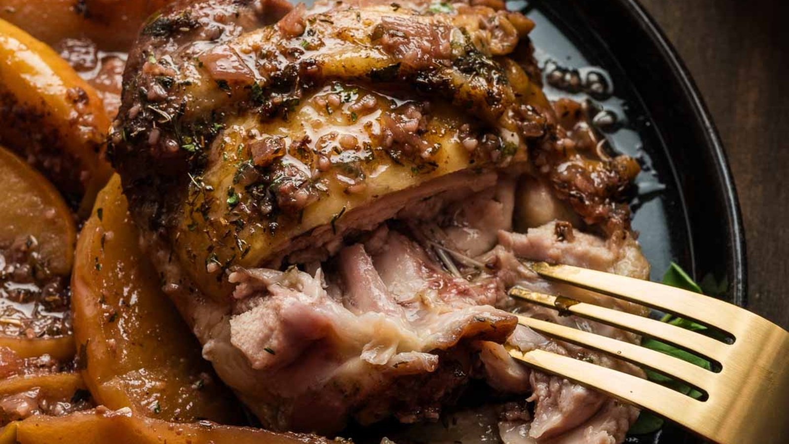 A close-up of a cooked piece of seasoned pork being pulled apart with a fork on a black plate, with sliced apples on the side—perfect for hearty dinners.