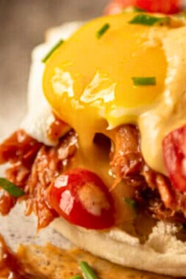 A close-up of a poached egg with runny yolk over shredded meat, sliced cherry tomatoes, sauce, and chives on a toasted English muffin.