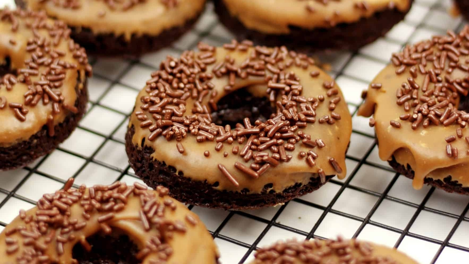 Chocolate donuts topped with tan glaze and chocolate sprinkles are arranged on a metal wire cooling rack.