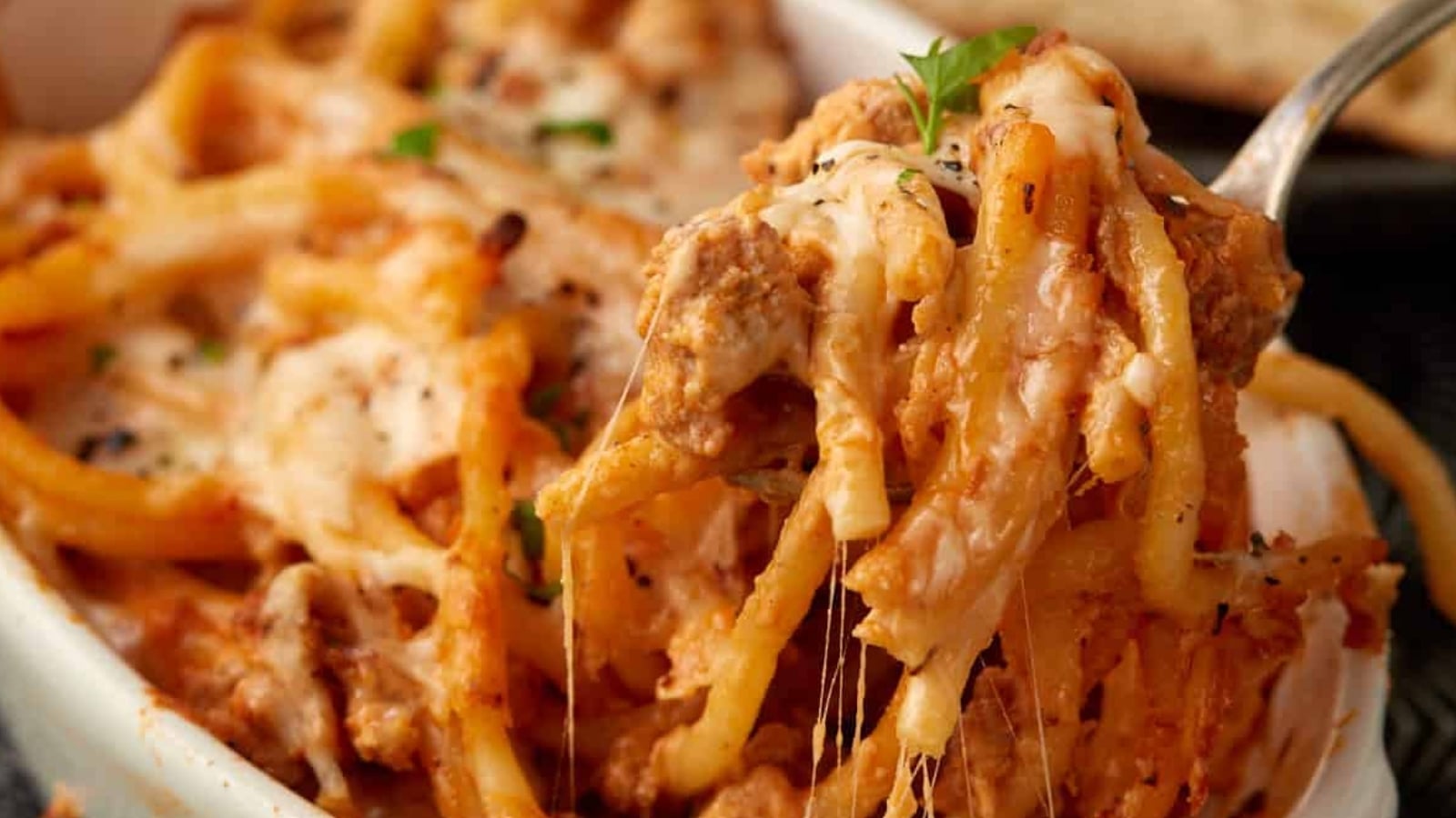 A close-up of baked spaghetti with melted cheese, tomato sauce, and ground meat being lifted with a fork from a casserole dish.