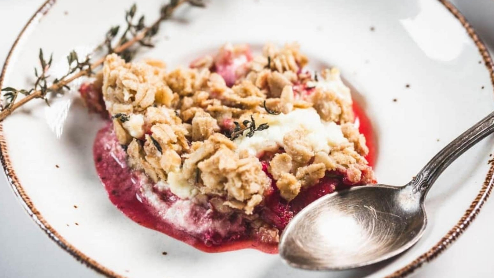 A serving of fruit crumble with a crumbly oat topping and a layer of red fruit filling, garnished with a sprig of thyme on a white plate with a brown rim, next to a silver spoon.