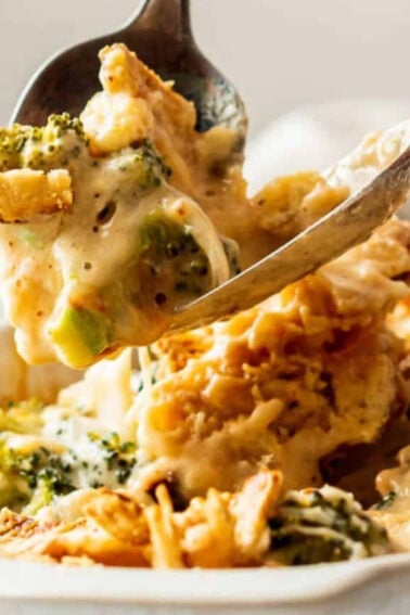 A serving spoon lifts a portion of cheesy broccoli and chicken casserole from a white baking dish.