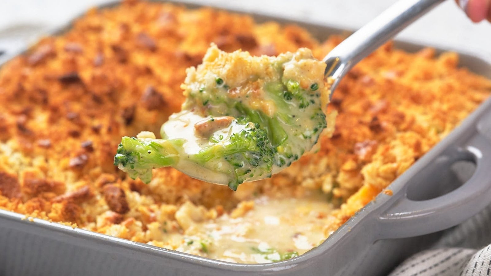 A close-up of a spoonful of broccoli casserole being lifted from a baking dish with a golden, crumbly topping.