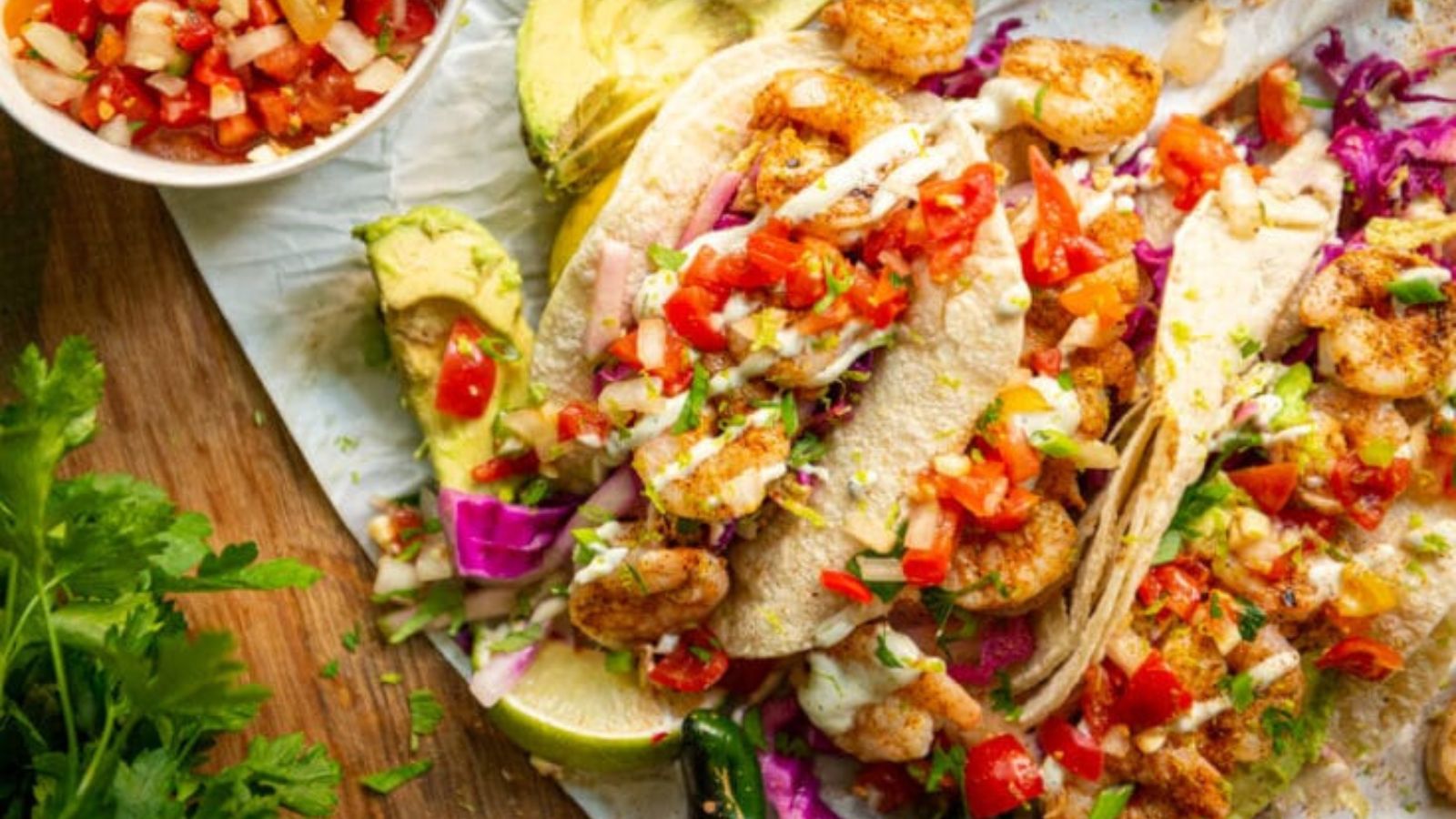 Three shrimp tacos topped with diced tomatoes, cabbage, and sauce are served with sliced avocado and a bowl of salsa on the side.