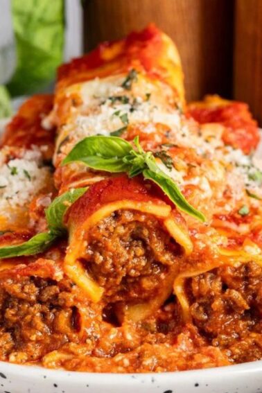 Three cannelloni pasta tubes filled with meat and tomato sauce, topped with grated cheese and fresh basil, served on a white plate with a fork.