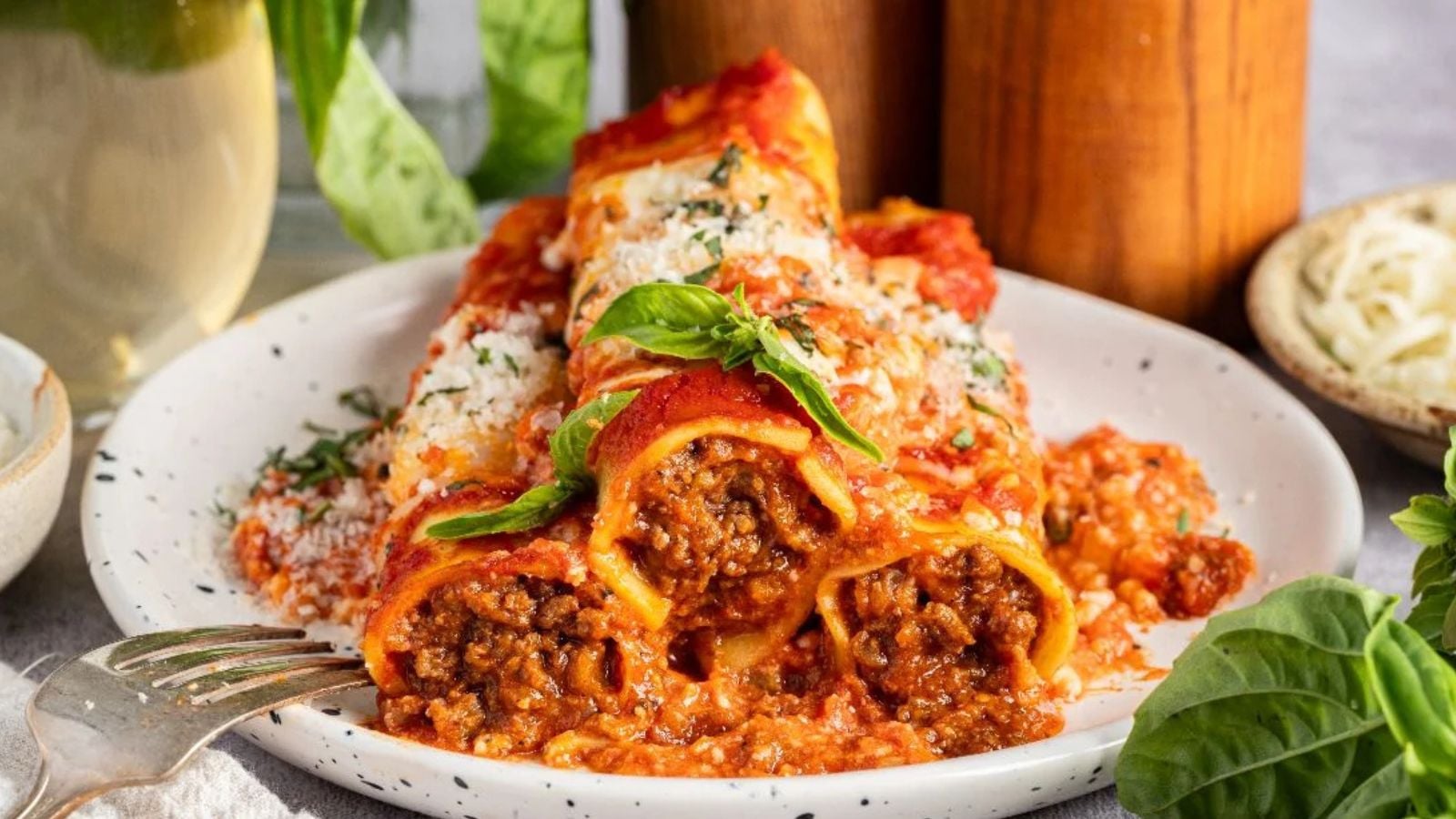 Three cannelloni pasta tubes filled with meat and tomato sauce, topped with grated cheese and fresh basil, served on a white plate with a fork.