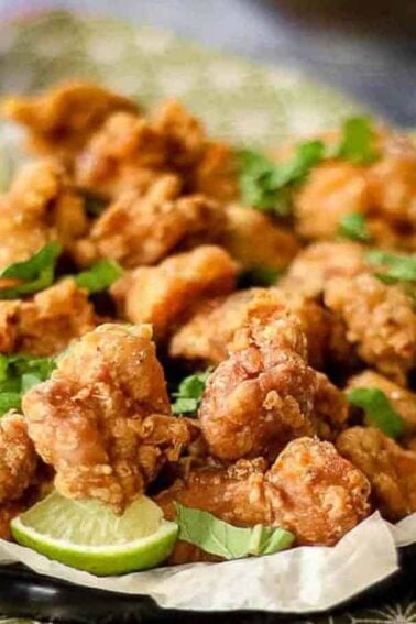 A plate of golden fried chicken pieces garnished with cilantro and lime wedges, served with dipping sauces in small bowls.